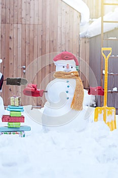 Smiling snowman in knitted mittens, scarf and red cap with gifts and shovel  in the back yard fence  background