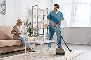 smiling social worker cleaning carpet with vacuum cleaner and looking at senior woman photo