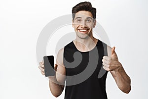 Smiling sportsman in activewear showing smartphone screen and thumb up, standing over white background