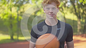 Smiling sportsman posing in sunlight on outdoor basketball court. Portrait of young confident redhead man holding ball