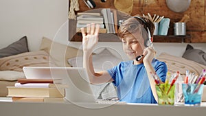 Smiling student boy studying at home with remote school connected to computer on the web, raise his hand to answer questions or