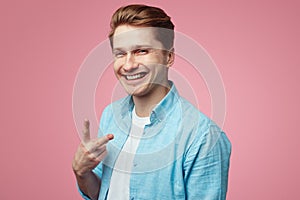 Smiling student wearing blue shirt and is gesturing vsign over pink wall