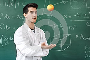 Smiling student with white coat throwing an apple up, on green chalkboard with equations background.