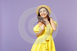 Smiling stunning young woman in yellow dress, summer hat standing, looking aside  on pastel violet wall