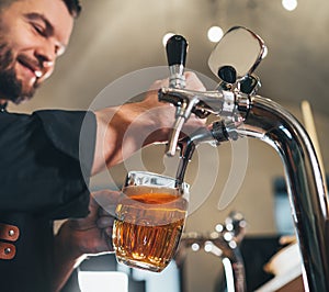 Smiling stylish bearded barman dressed black uniform with an apron tapping fresh lager beer into glass mug at bar counter. photo