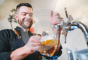 Smiling stylish bearded barman dressed black uniform with an apron tapping fresh lager beer into pilsner glass mug at bar counter