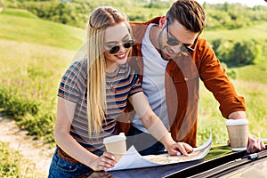 smiling stylish couple of tourists with paper cups of coffee looking for destination on map near car in rural