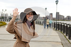 Smiling stylish girl in a coat with belt and big hat waving HELLO. The concept of welcome, friendliness, fun