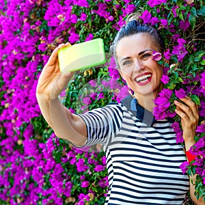 Smiling stylish woman near flowers bed taking selfie with phone