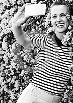 Smiling stylish woman near flowers bed taking selfie with phone