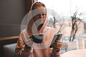 Smiling stylish young girl wear hood drink coffee holding phone in cafe close up.