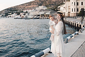 Smiling stylish young woman hold baby girl 1 year old wear white trendy dress walk on quayside over sea shore outdoors together.