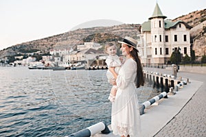 Smiling stylish young woman hold baby girl 1 year old wear white trendy dress walk on quayside over sea shore outdoors together.