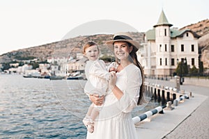 Smiling stylish young woman hold baby girl 1 year old wear white trendy dress walk on quayside over sea shore outdoors together.