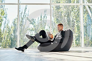 Smiling successful businessman in elegant suite using tablet sitting on bean bag chair