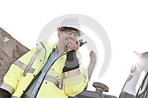 Smiling supervisor using walkie-talkie at construction site against clear sky