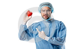 Smiling surgeon in blue medical uniform and medical mask and pointing at toy heart