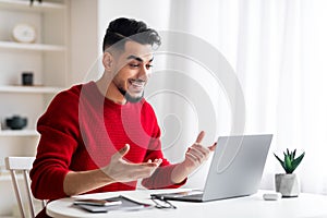 Smiling surprised young middle eastern male with beard looks at computer and gesticulate at workplace in home