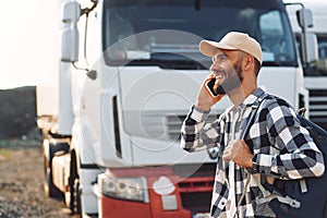 Smiling, talking by phone. Young truck driver is with his vehicle at daytime