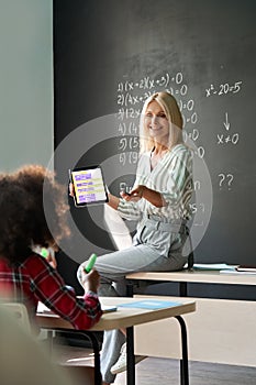 Smiling teacher having maths class sitting on desk holding tablet device.