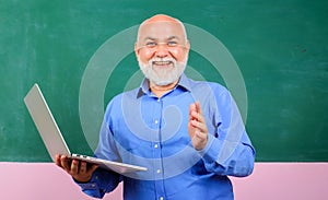 Smiling teacher with laptop in front of blackboard at classroom. Teacher giving lesson to students. E-learning at
