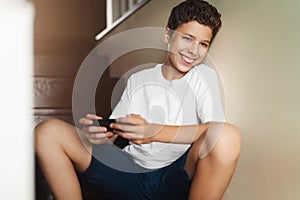Smiling teen boy sitting at home on steps, using smartphone. Guy in white T-shirt is chatting, playing games on his cell phone