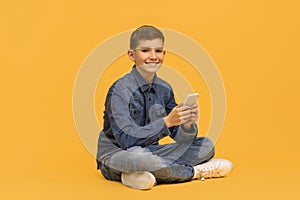 Smiling teen boy with smartphone in hands sitting cross-legged on yellow background