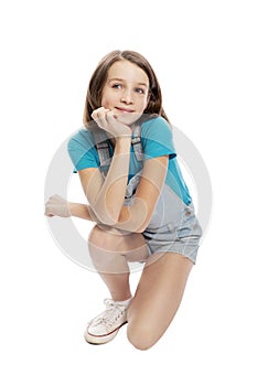 Smiling teen girl in denim overalls squats. Isolated on a white background.