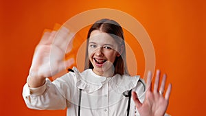 Smiling teen girl waving hello greeting against orange background in studio