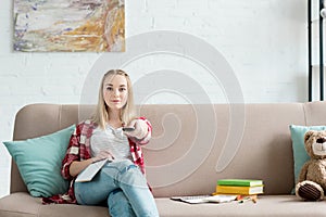 smiling teen student girl with remote control watching tv while sitting on couch with book