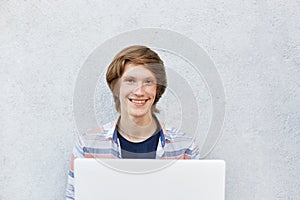 Smiling teenage boy with attractive appearance sitting over white background using laptop for watching films or playing games. Col