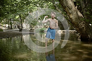 Smiling teenage boy posing like bodybuilder in shallow water in forest in mediterranean country in summer time, sunny relaxed mood