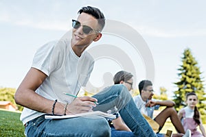 smiling teenage boy in sunglasses taking notes and looking away while studying with friends