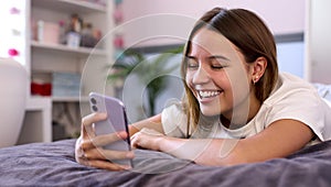 Smiling Teenage Girl Lying On Bed At Home Looking At Mobile Phone