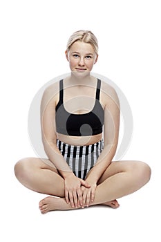 A smiling teenage girl in a sports swimsuit sits on the floor. A cute blonde with freckles on her face in a black top and striped