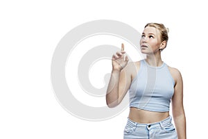 A smiling teenage girl stands and points her index finger up. Cute blonde in jeans and a blue top. Isolated on a white background