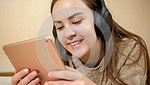 Smiling teenage girl with tablet computer lying on bed and listening music in headphones