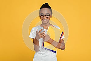 Smiling teenager african american girl pupil in glasses and casual holds books and reads message on smartphone
