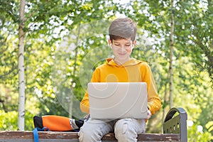 Smiling teenager boy working on laptop. Holding and using a laptop for networking on a sunny spring day, outdoors.