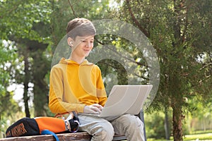 Smiling teenager boy working on laptop. Holding and using a laptop for networking on a sunny spring day, outdoors.