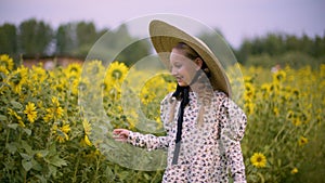 Smiling teenager girl in hat touching sunflowers on summer walk at rustic field. Happy young girl laughing on walk at