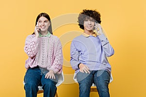 Smiling teenagers talking on cellphones on chairs on yellow background.