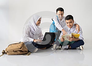 smiling three teenagers in junior high school uniforms sitting on the floor studying together using a laptop and a book