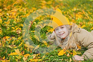 smiling tlittle preschool girl with blond hair in a yellow hat lies on the grass with autumn leaves portrait of a