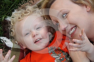 Smiling toddler boy cuddles outdoors on blanket with pretty mother. photo