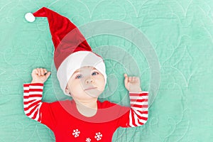 Smiling toddler boy lying on green coverlet. Christmas baby wearing big red Santa hat looking at camera. New Year