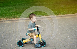 A smiling toddler boy of two or three years old rides a bicycle or balance bike in a city park on a sunny summer day.