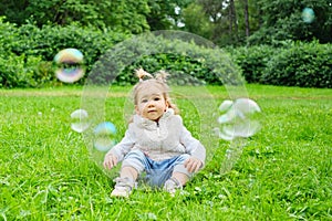 A smiling toddler sits on the grass in a summer park and looks at soap bubbles