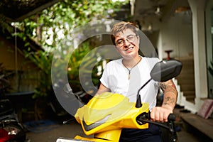 Smiling transgender individual on yellow scooter enjoys tropical setting. Person ready for ride, embracing freedom, warm