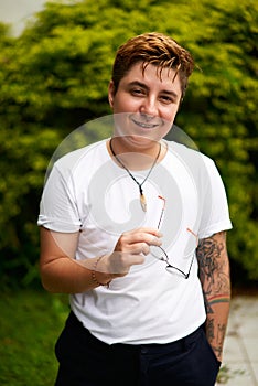 Smiling transgender man outdoors in casual attire, confidence evident in stance with glasses in hand, tattoo visible on